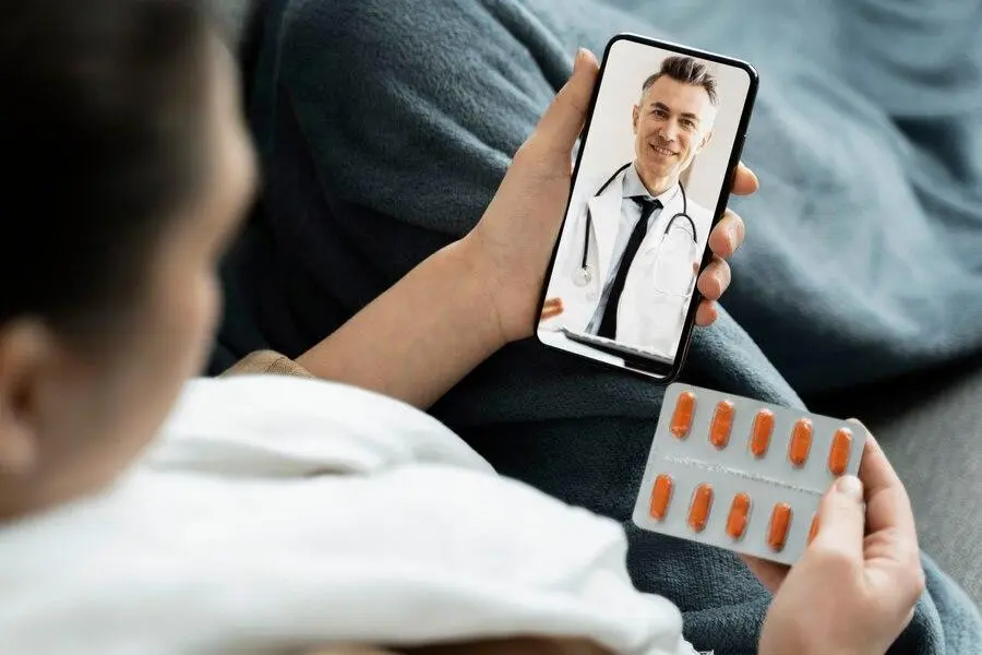 Person holding a smartphone with a male doctor on the screen, suggesting a telemedicine consultation, while also holding a blister pack of pills.