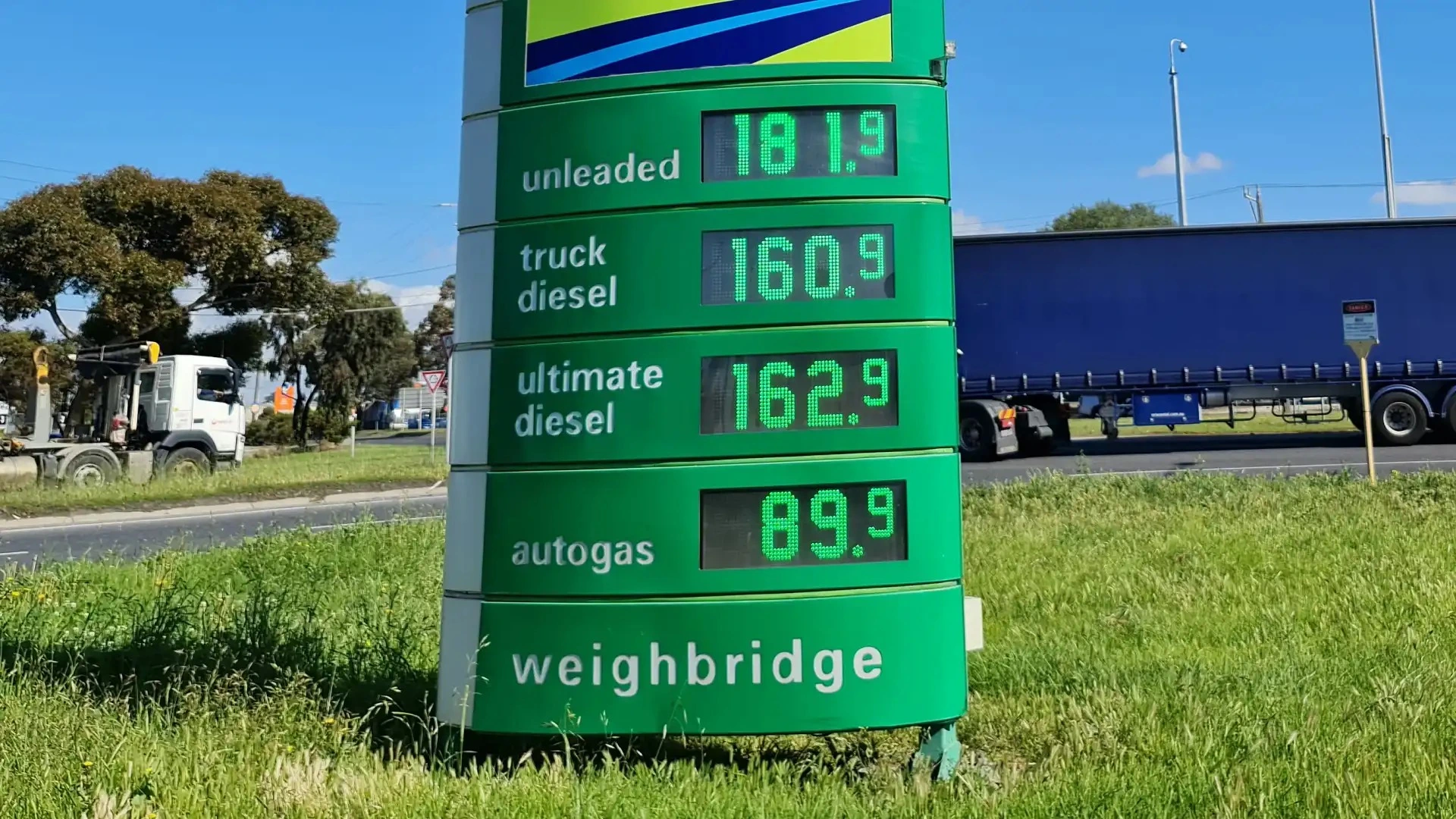 Green gas station sign displaying prices for various types of fuel including Unleaded, Truck Diesel, Ultimate Diesel, and Autogas, with a blue truck passing by in the background